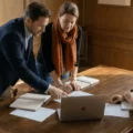 Three colleagues collaborate around a wooden table: two leaning over papers while a third sits with a laptop and notebook open nearby, in a meeting room.