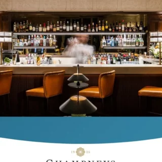 Bar interior with liquor shelves and four orange leather stools along a white counter; a blurred bartender in the center.