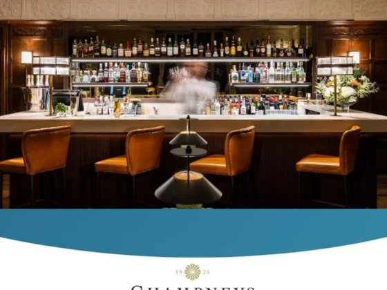 Bar interior with liquor shelves and four orange leather stools along a white counter; a blurred bartender in the center.