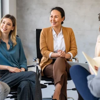 Five professionals seated in a circle in a bright conference room, smiling and chatting during a team discussion.