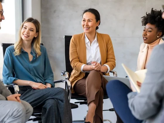 Five professionals seated in a circle in a bright conference room, smiling and chatting during a team discussion.