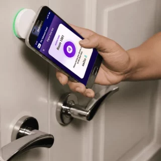 Person holding a smartphone near a smart door lock mounted on a door; app screen visible in the phone display