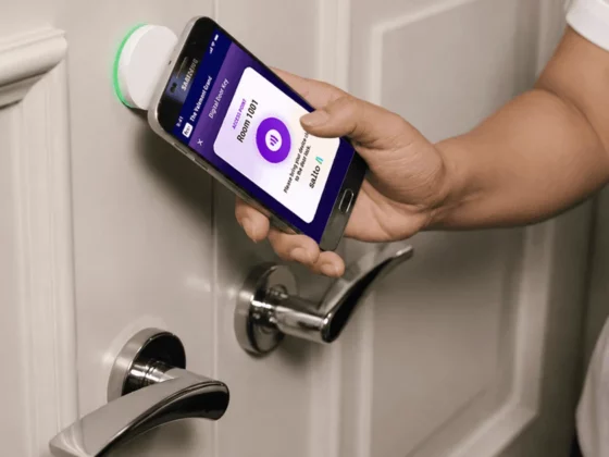Person holding a smartphone near a smart door lock mounted on a door; app screen visible in the phone display