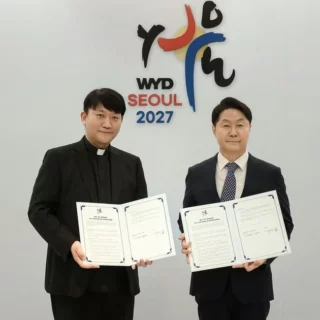 Two men in formal attire hold open documents and pose for a signing ceremony in front of a WYD Seoul 2027 backdrop, flanked by Vatican and South Korean flags.