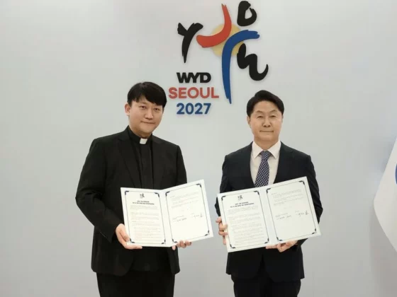 Two men in formal attire hold open documents and pose for a signing ceremony in front of a WYD Seoul 2027 backdrop, flanked by Vatican and South Korean flags.