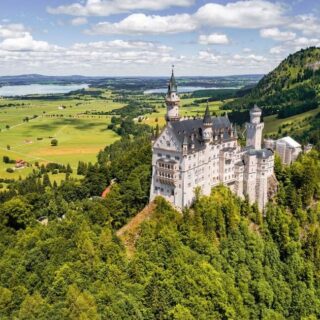 Fairy-tale castle perched on a hillside amid dense forest with green valley and lake in distance, under a blue sky with clouds.