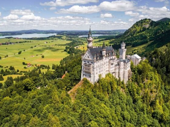 Fairy-tale castle perched on a hillside amid dense forest with green valley and lake in distance, under a blue sky with clouds.