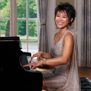 Smiling woman in a silver sequined dress plays a glossy Steinway & Sons grand piano in a bright room with large windows behind her.