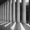 Row of tall columns along a covered walkway, casting long diagonal shadows on the floor in black and white.