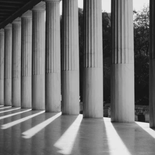 Row of tall columns along a covered walkway, casting long diagonal shadows on the floor in black and white.