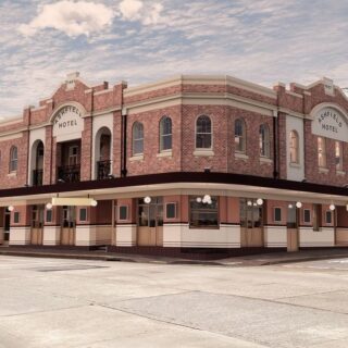 Corner view of a brick hotel with arched windows and a long beige ground-floor storefront under a row of string lights, on a city street.