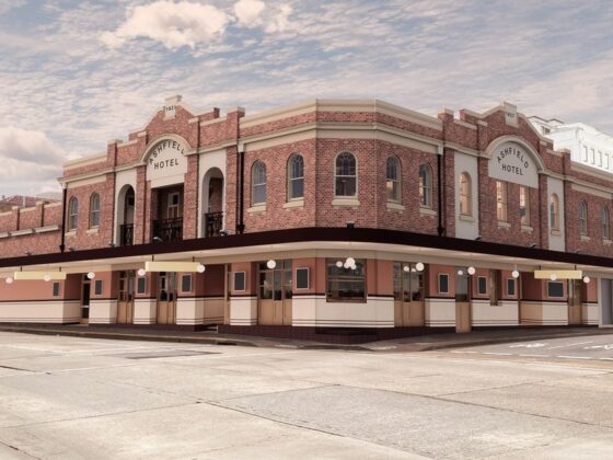 Corner view of a brick hotel with arched windows and a long beige ground-floor storefront under a row of string lights, on a city street.