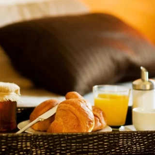 Bedside breakfast tray on a bed with croissants, orange juice, and small condiments.