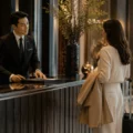 Hotel concierge in a suit checks in a guest at a dark wood front desk in a luxury lobby with a rolling suitcase nearby.
