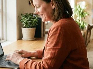 Woman typing on a laptop at a wooden table by a sunlit window, with a potted plant beside her.
