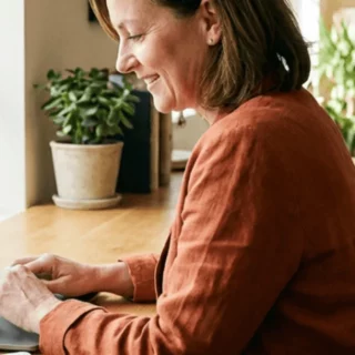 Woman typing on a laptop at a wooden table by a sunlit window, with a potted plant beside her.