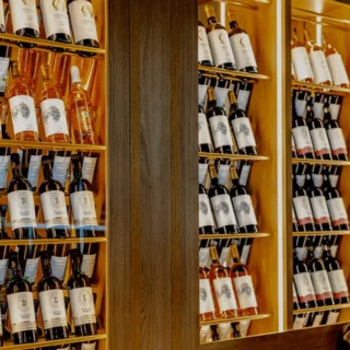 Wine bottles on illuminated wooden shelves in a bar, with a vintage clock at the bottom right corner.