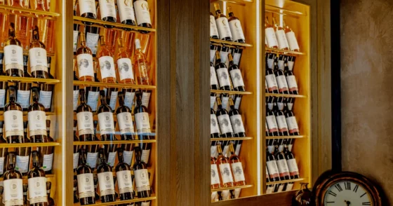 Wine bottles on illuminated wooden shelves in a bar, with a vintage clock at the bottom right corner.