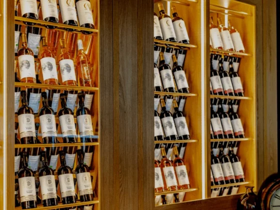 Wine bottles on illuminated wooden shelves in a bar, with a vintage clock at the bottom right corner.