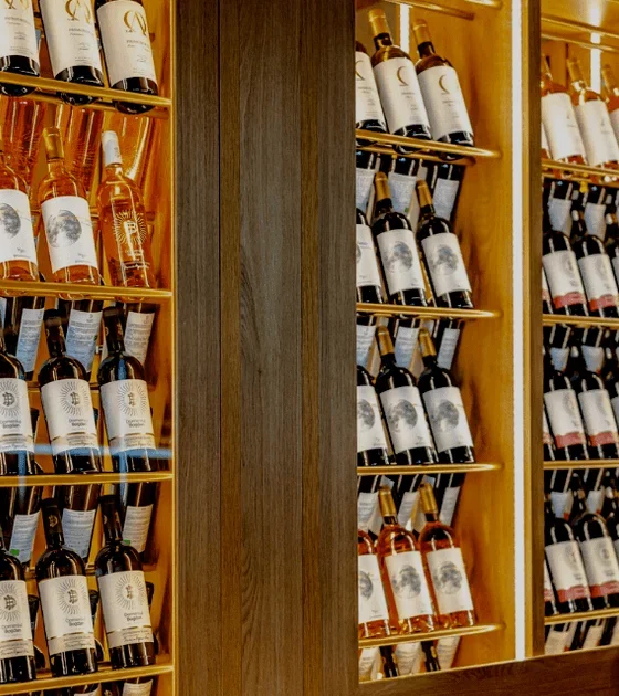 Wine bottles on illuminated wooden shelves in a bar, with a vintage clock at the bottom right corner.
