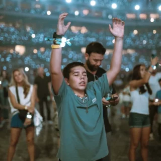 Boy in a teal shirt raises his arms in a crowded, dimly lit concert, wearing colorful wristbands as lights glow behind him.