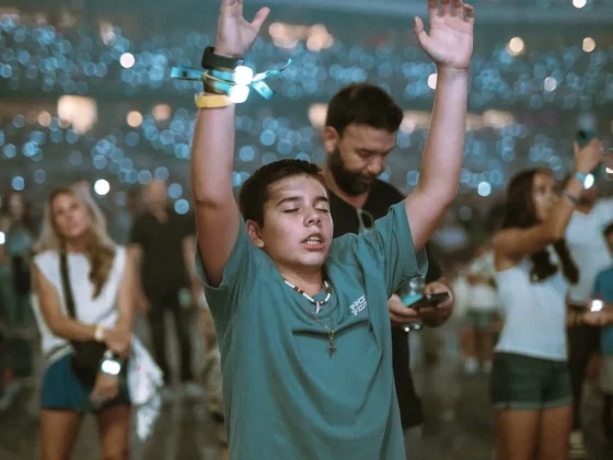 Boy in a teal shirt raises his arms in a crowded, dimly lit concert, wearing colorful wristbands as lights glow behind him.