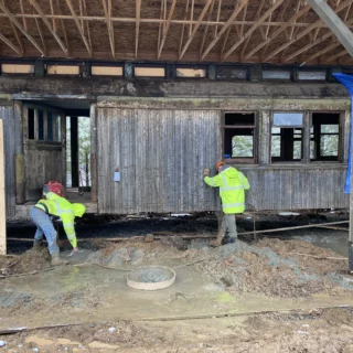 Construction workers in high-visibility jackets stand and crouch in a muddy construction site inside a weathered wooden building with exposed roof trusses and a blue tarp on the right side.