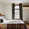 Cozy bedroom with a queen bed, plaid throw, wicker headboard, two lamps, and a wooden dresser with a round mirror.