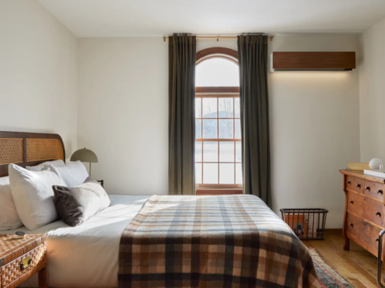 Cozy bedroom with a queen bed, plaid throw, wicker headboard, two lamps, and a wooden dresser with a round mirror.