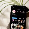 Close-up of a smartphone displaying an Instagram profile, with the circular logo and follower counts, inside a white basket next to a green plant.