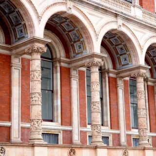Row of decorative arches on a red brick building, supported by carved stone columns with intricate detailing above large windows.