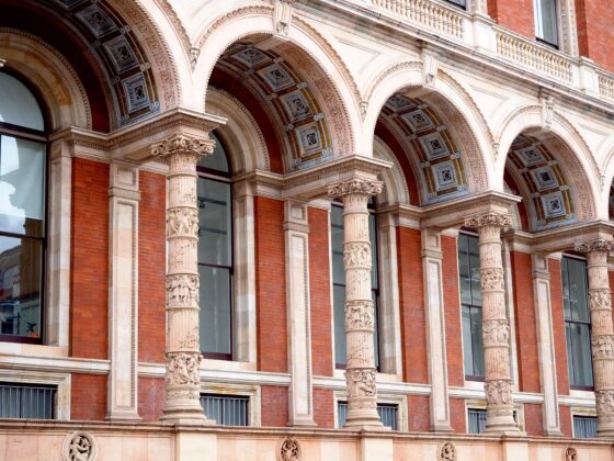 Row of decorative arches on a red brick building, supported by carved stone columns with intricate detailing above large windows.