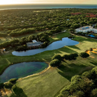Aerial view of a golf course with ponds, greens, and a clubhouse nestled among trees at sunset.