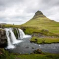Wide landscape featuring a cone-shaped mountain with a small waterfall cascading into a river, under a cloudy sky.