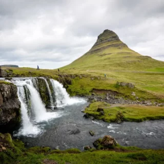 Wide landscape featuring a cone-shaped mountain with a small waterfall cascading into a river, under a cloudy sky.
