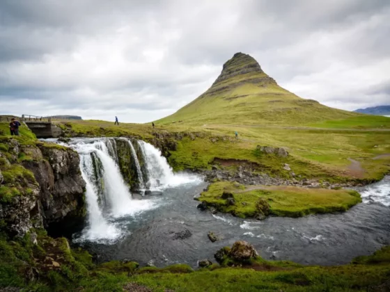 Wide landscape featuring a cone-shaped mountain with a small waterfall cascading into a river, under a cloudy sky.