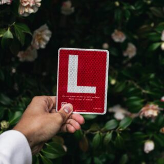 Hand holding a red reflective square sign with a white L-shaped corner against a backdrop of green leaves and pale pink flowers.
