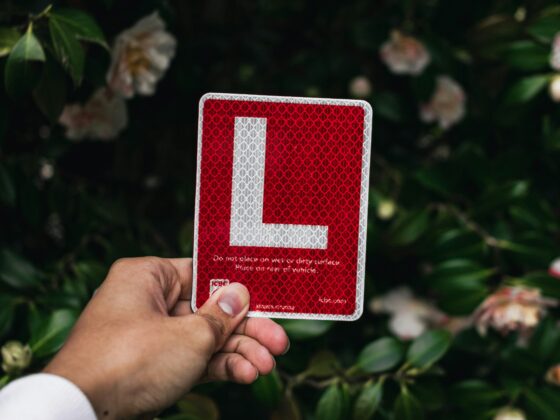 Hand holding a red reflective square sign with a white L-shaped corner against a backdrop of green leaves and pale pink flowers.
