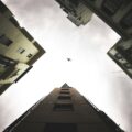 Worm's-eye view of tall apartment buildings forming a square around a cloudy sky, with a small airplane silhouetted in the center.