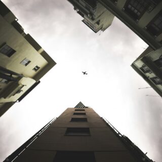 Worm's-eye view of tall apartment buildings forming a square around a cloudy sky, with a small airplane silhouetted in the center.