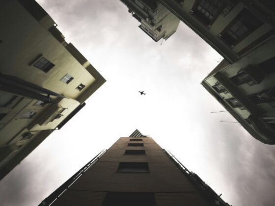 Worm's-eye view of tall apartment buildings forming a square around a cloudy sky, with a small airplane silhouetted in the center.