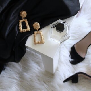 Gold textured dangle earrings and a clear square perfume bottle on a white surface next to a black fabric and a black-strap heeled sandal in the background.