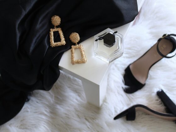 Gold textured dangle earrings and a clear square perfume bottle on a white surface next to a black fabric and a black-strap heeled sandal in the background.
