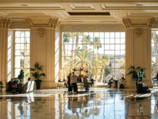 Sunlit hotel lobby with tall windows, ornate columns, and plants; people gather around tables with a floral centerpiece nearby.