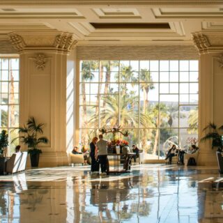 Sunlit hotel lobby with tall windows, ornate columns, and plants; people gather around tables with a floral centerpiece nearby.
