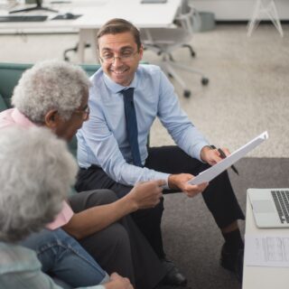 Three colleagues—an older couple and a younger man in a blue shirt and tie—review documents on a white table with a laptop nearby in a bright office.