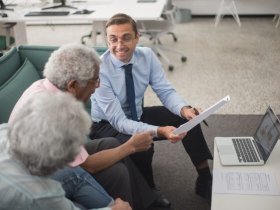 Three colleagues—an older couple and a younger man in a blue shirt and tie—review documents on a white table with a laptop nearby in a bright office.