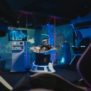 Person wearing a VR headset sits in a futuristic capsule ride under blue and pink neon lights in an arcade setting.