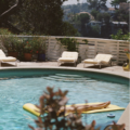 Person sunbathing on a yellow float in a swimming pool, with lounge chairs along a white fence behind them.