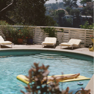 Person sunbathing on a yellow float in a swimming pool, with lounge chairs along a white fence behind them.